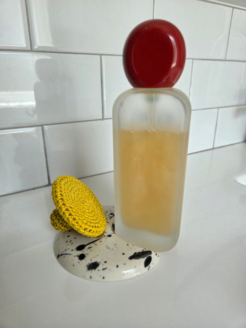 Yellow Mihakka exfoliation tool sitting on moon shaped rest next to bottle of Alia Jouj (red cap, golden color cleanser); background is white tile