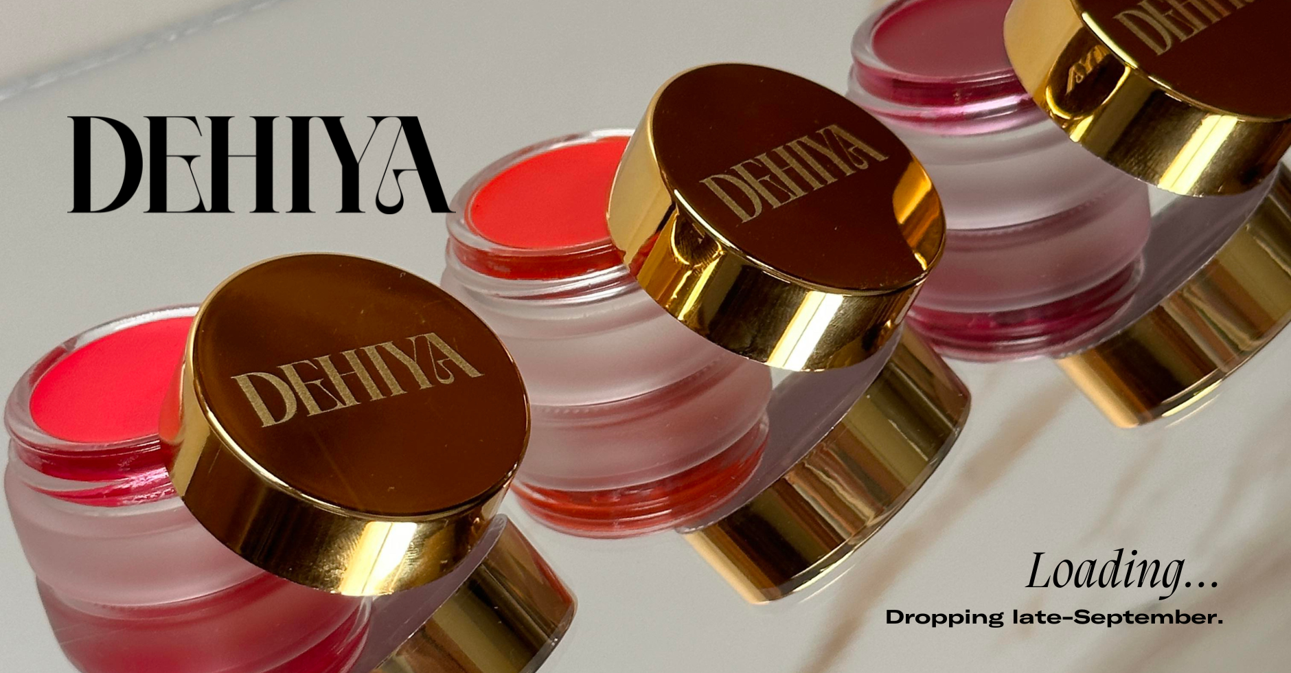 DEHIYA Logo, Text: Loading...dropping late-September, trio of lip and cheek pots open with gold caps, colors are pink, orange-red, dark pink